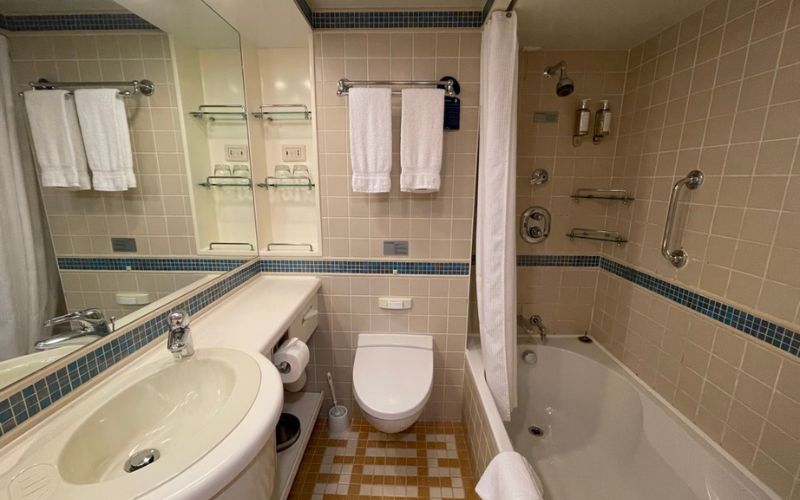 A clean and well-lit bathroom with beige tiles and blue accents, featuring a sink, toilet, and a bathtub with a shower curtain. White towels hang neatly on a rack above the toilet, and various toiletries are organized on shelves and in dispensers.