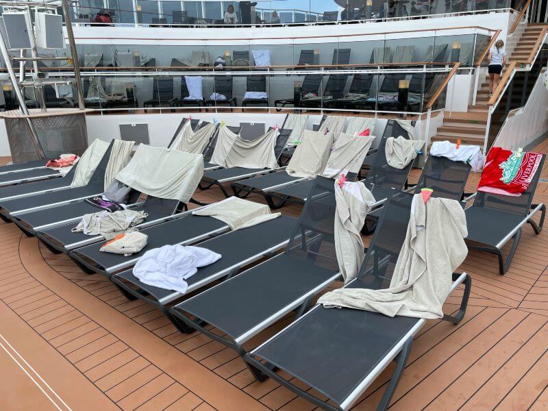 Several sun loungers on the deck of a cruise ship are covered with towels, indicating they have been reserved by passengers. The loungers are arranged in rows, with some towels draped and others neatly folded, set against the backdrop of multiple deck levels and glass railings.