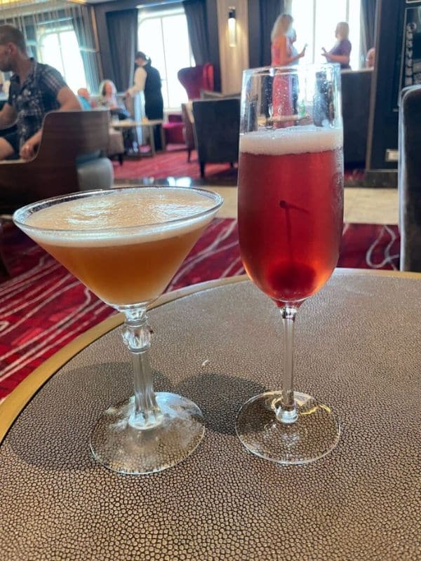 Two elegantly presented cocktails on a textured table, with a vibrant cruise ship lounge atmosphere in the background.