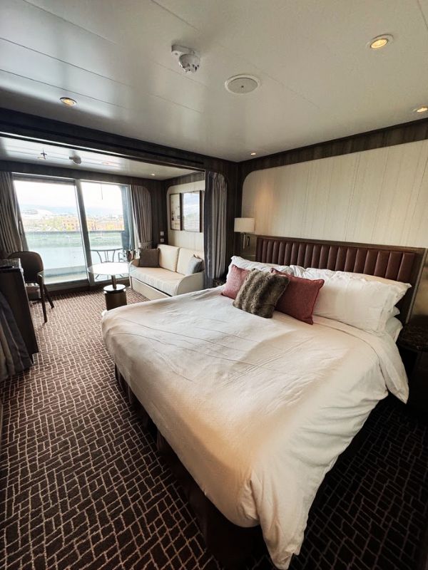 The elegant Verandah Suite on the Seabourn Venture, designed to provide a luxurious and comfortable accommodation experience. The suite features a large, plush king-sized bed with stylish bedding and decorative pillows. A cozy seating area near the window includes a sofa and a coffee table, perfect for relaxing and enjoying the view. The room is tastefully decorated with neutral tones, textured walls, and modern furnishings, creating a sophisticated ambiance. Large sliding glass doors lead to a private verandah, offering guests beautiful views of the port and the opportunity to enjoy the sea breeze.