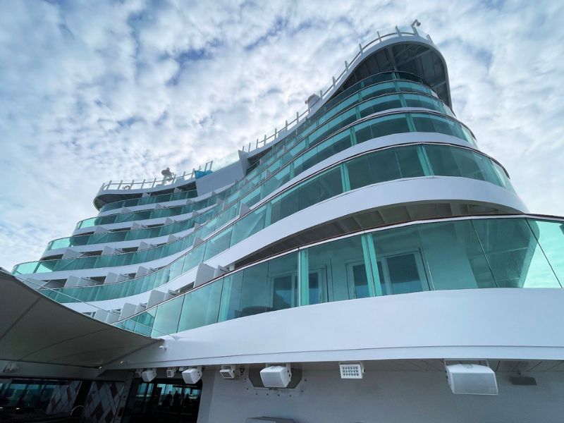 The aft of a cruise ship showcasing the tiered deck levels with glass balustrades, under a soft blue sky with scattered clouds.