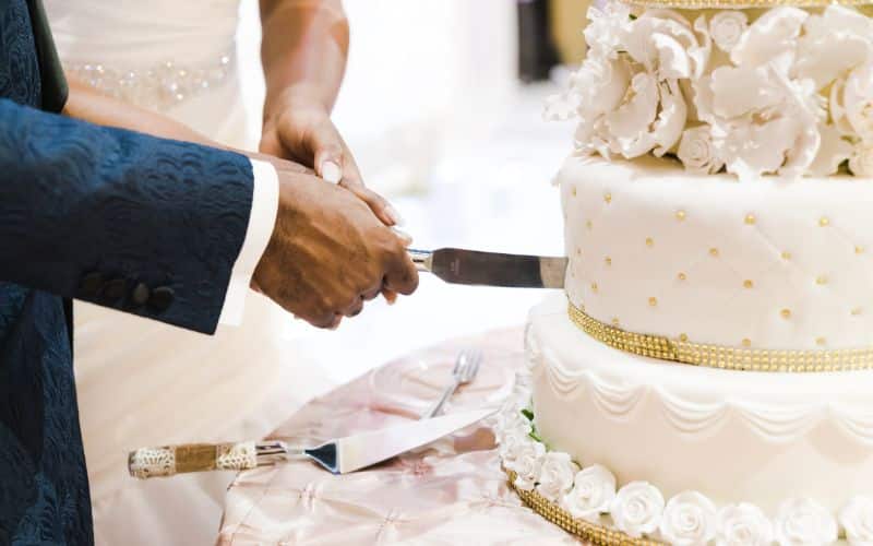 A newlywed couple jointly holds a knife and cuts into a lavish multi-tiered wedding cake adorned with white floral details and golden accents on a decorated table.