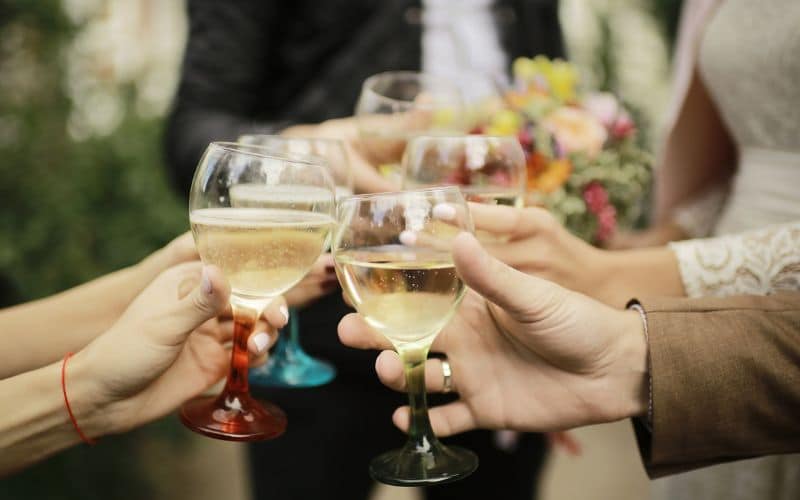Guests at a wedding celebration toasting with glasses of white wine, featuring close-up views of hands holding colorful glass stems. The backdrop is subtly blurred, highlighting the festive mood and the floral details from the event.