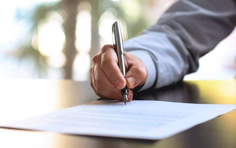 Close-up of a person's hand signing a document with a pen, focused on the action of writing with a blurred background featuring natural light and greenery. The scene conveys a formal, significant moment, likely related to legal or official paperwork.