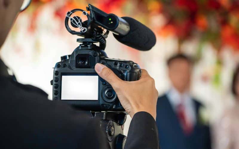 A photographer is capturing a wedding ceremony with a professional camera, focusing on the couple in the background, who are slightly blurred. The camera's microphone and a vibrant floral backdrop are also visible.