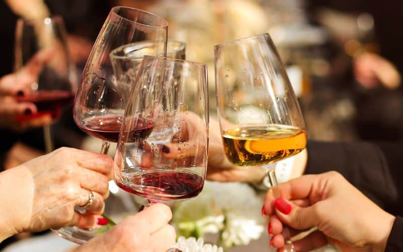 Close-up of hands clinking wine glasses filled with red and white wine at a festive gathering. The glasses are elegantly shaped, capturing the light and reflections of the joyous event.