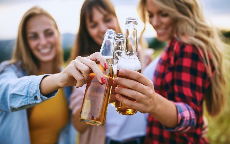 Three smiling women are toasting with beer bottles outdoors, enjoying a casual gathering. They are dressed in casual attire, including a red plaid shirt and light jackets, with a blurred scenic background.