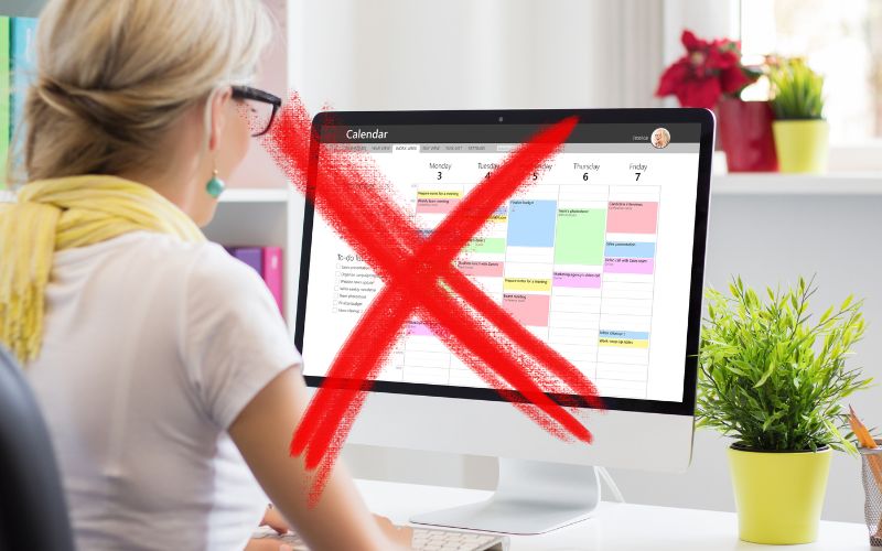 The image depicts a woman viewing a computer screen with a digital calendar filled with colorful events, overlaid with a large red 'X'. This suggests either an error or cancellation related to the scheduled events. The office environment is bright and modern, featuring indoor plants and a clean, organized desk setup.