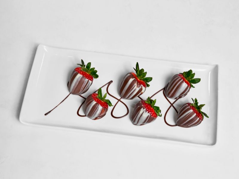 Five fresh strawberries offered by NCL onboard, dipped in dark chocolate and drizzled with white chocolate, elegantly presented on a rectangular white plate against a minimalist background.